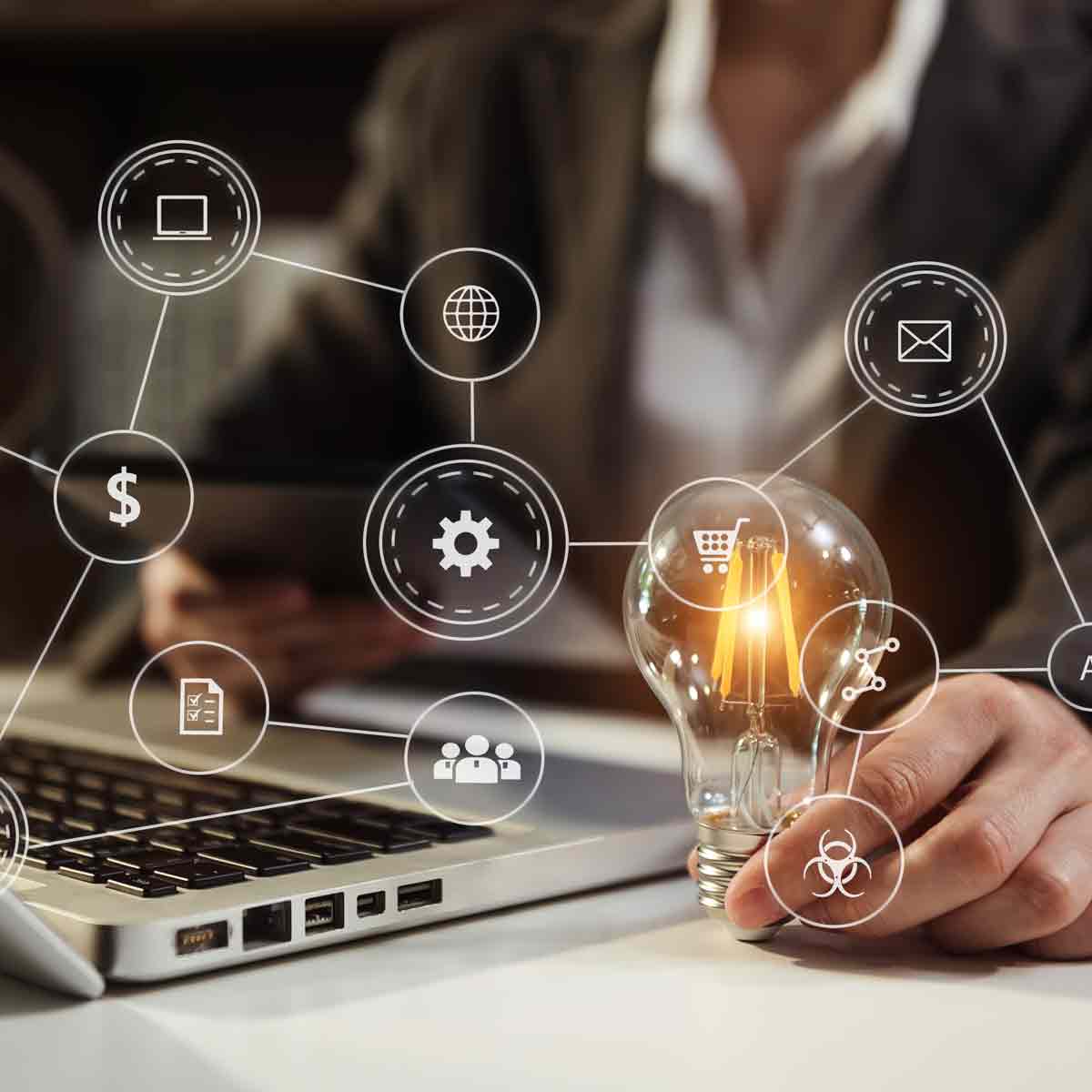 Mano che tiene una lampadina accesa accanto a un laptop con icone di tecnologia e connessione sovrapposte.